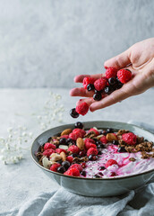 Hand throws berries in bowl with yogurt, granola, muesli, seeds, frozen raspberries, nuts on light background. Healthy food, detox smoothy, vegetarian nutrition, close up
