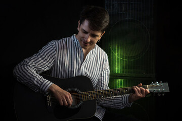 Photo of man sitting and playing acoustic guitar in recording studio on dark background with light...