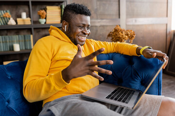 Joyful black businessman sitting on sofa at home, looking at the computer screen, emotionally...