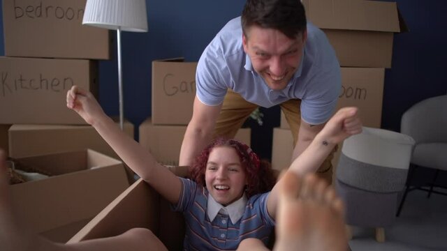 Close portrait of a teenage girl with bare feet sits in a cardboard box while her happy father rolls her around a new house