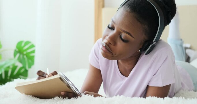 Young woman writing to do list in notepad while listening music on headphones in bed at home