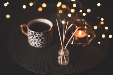 Cup of black fresh coffee with tasty dessert and glowing candle, glass bottle with wood sticks over Christmas lights close up. Cozy home atmosphere. Winter holiday season. Celebration.