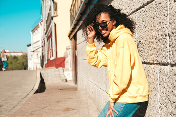 Beautiful black woman with afro curls hairstyle.Smiling hipster model in yellow hoodie. Sexy carefree female posing on the street background in sunglasses. Cheerful and happy