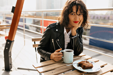 Charming attractive young woman with short black curly hair and red lips drinking coffee with dessert on open air terrace in the morning
