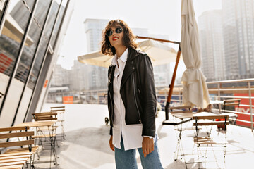 Attractive stylish girl with short hairstyle wearing white shirt and black jacket posing on the summer terrace in sunlight