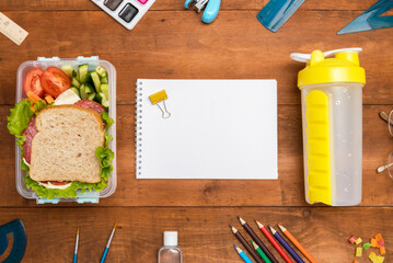 A school lunch box with a sandwich, vegetables, water, fruit and a notebook on a wooden background. School supplies,and food. Flatly.The concept of education and nutrition.Mock up.