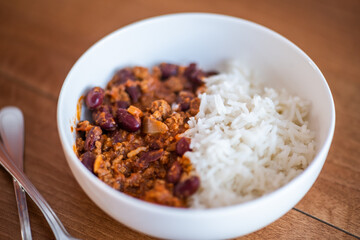Close up and selective focus of meat chilli concarne and white rice in a white bowl on a wooden surface with intentional shallow depth of field and bokeh