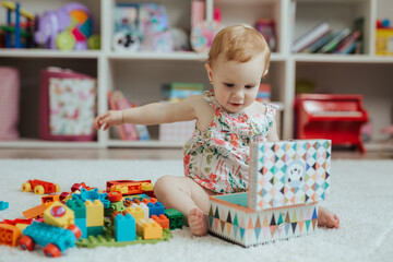 Fototapeta na wymiar Little curious baby girl playing on a playground in a bright sunny room. Learning and playing colorful educational blocks game and puzzle