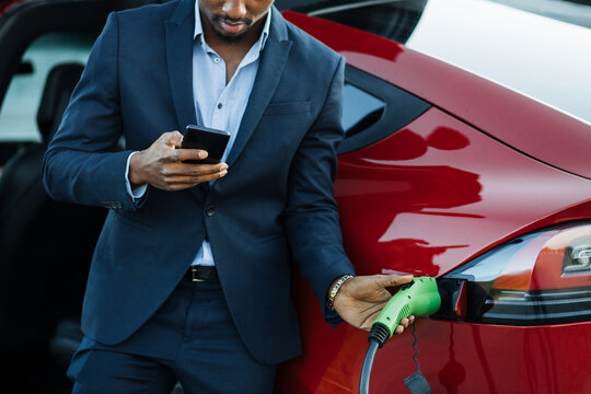 Close Up Of African Businessman Unplugging Charging Cable From Electric Car While Using Modern Smartphone. Concept Of Lifestyles, Ecology Vehicle And Busy People.