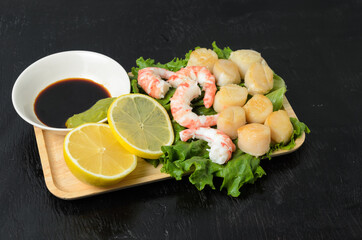 Fried scallop with boiled shrimp tail on a salad leaf with soy sauce and wasabi on a dark background in a wooden plate.