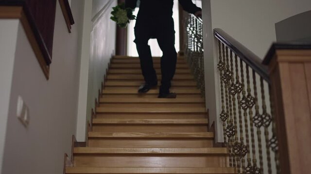 Hurrying groom in elegant suit running downstairs with bouquet of flowers. Young stylish man in wedding costume late for marriage ceremony