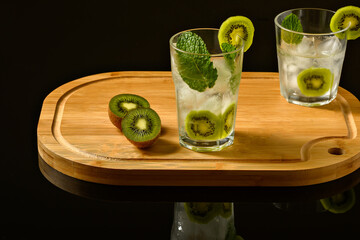 two glasses with iced drink decorated with kiwi mint and ice, on an insulated wooden board on a black background