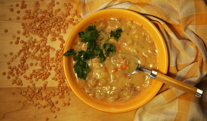 split pea soup in yellow plate, decorated with green parsley, split peas and linen cloth next to, top view, wooden table