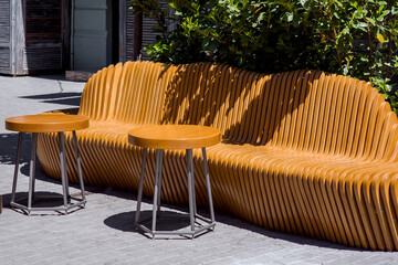 wooden art bench with creative design of brown wooden planks with round tables on the pedestrian sidewalk of a city street lit by sunlight nobody.