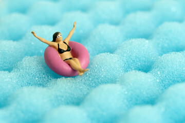 Miniature people toy figure photography. Girl wearing black sunglass swimming with rubber tube ring on wavy ocean.
