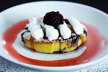 Sweet, juicy muffin with berries, sprinkled with vanilla sugar and whipped cream on top, with blackberries on a white and black plate