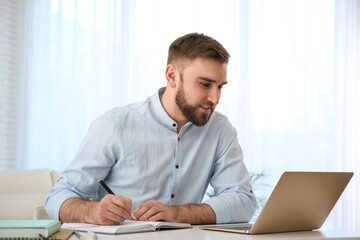 Fototapeta premium Young man taking notes during online webinar at table indoors