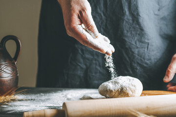 Male hands pour flour on the dough next to clay pot and oil bottle and rolling pin on dark table, while cooking