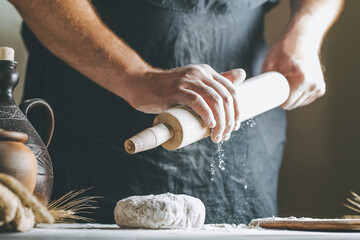 Male hands pour flour on a rolling pin next to dough, clay pot, oil bottle and rolling pin on dark table, while cooking