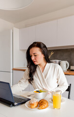 Young woman in comfortable white robe works on laptop while sitting at table the kitchen. Work at home for breakfast.