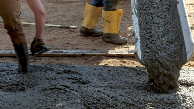 New cement concrete pours out of the moveable chute of a mixer truck, and a construction worker in rubber boots moves the chute as needed.
