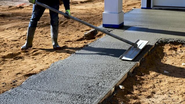 A bull float is used on new cement concrete by a worker to bring liquid to the surface, and to level and smooth the new sidewalk at a construction job site.