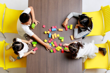 Top view shot group of Asian little cute intelligent kindergarten schoolboy and schoolgirls sitting on yellow chairs playing colorful brick toy together on wooden table at pre elementary school