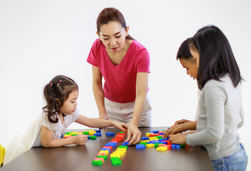 Group of Asian little cute intelligent kindergarten schoolboy and schoolgirls sitting on yellow chairs playing colorful brick toy together on wooden table at pre elementary school