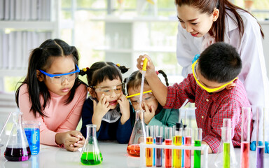 Asian little boy drop reagent sample by glass dropper into colorful solution flask while young girls wears safety glasses and teacher in white lab coat look at him do experiment in school laboratory.