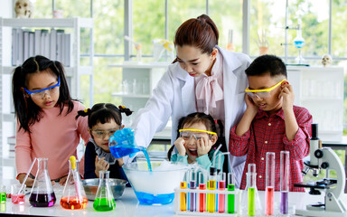 Group of Asian elementary little boy and girls wear safety goggles excited and looking at female science teacher in lab coat pouring blue reagent from beaker into glass bowl has smoke reaction
