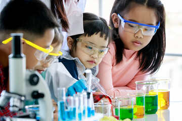 Asian little curious preschool girl focusing observing at teacher pouring red reagent from glass beaker do chemical experiment on laboratory table with microscope and equipment with young friends