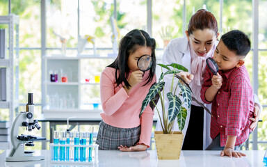 Obraz premium Asian female botany teacher in white lab coat teach botanical knowledge to primary boy and girl students using magnifying glass study plant leaf on laboratory table full with microscope test tubes