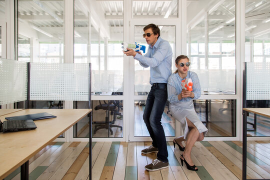 Man and woman in formal outfits standing back to back and aiming toy guns while having fun in office