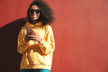 Beautiful black woman with afro curls hairstyle.Smiling model in yellow hoodie. Sexy carefree female posing on the street near red wall in sunglasses. Looking at smartphone screen, using apps