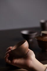 Cropped shot of a hand holding brown squat teabowl. Cup with ribbed textured surface is made in classic Japanese style. Equipment for tea ceremony is located in the background. 