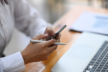 Close up view businesswoman holding pen and using smart phone at office desk.