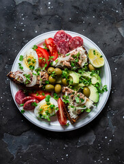 Large snack, appetizers, tapas plate - salami, mackerel cream cheese sandwiches, olives, avocado, tomatoes on a dark background, top view