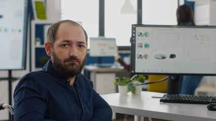 Fototapeta premium Close up of disabled invalid entrepreneur sitting in wheelchair looking at camera sad working in economic business company at computer prosessing financial information, graphs monitoring economy