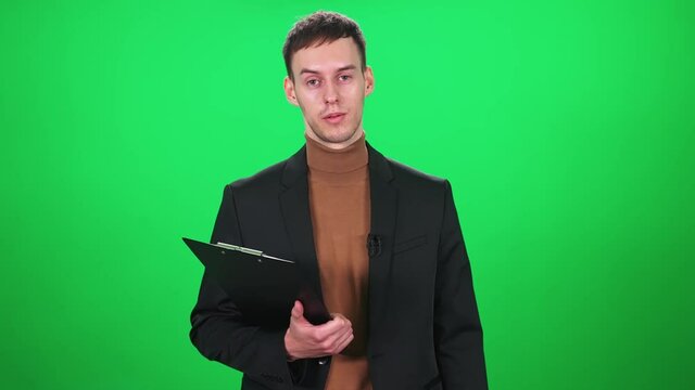 Man reporter in suit looks into the camera and speaks in lavalier microphone, holding a folder of documents in his hands, man on a green background, a template for TV news agencies, journalist at work