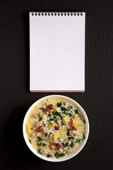 Homemade Zuppa Toscana with Kale and Bread in a white bowl, blank notepad on a black surface, top view. Overhead, from above, flat lay.
