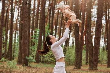 Profile of mother playing with baby girl, woman throwing up small daughter in air, happy family having fun outdoor, mom and child resting in forest, enjoying nature.