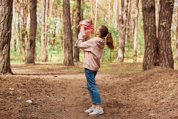 Full length outdoor shot of loving woman throwing up her infant daughter in air, mother and kid playing in forest, enjoying weekend in nature, family wearing casual clothing.