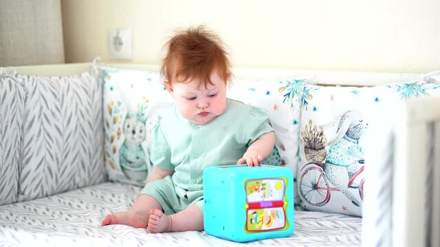 A baby with red hair sits in a crib and plays with a musical toy