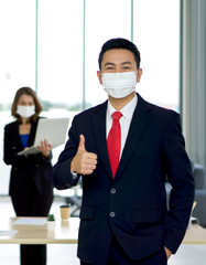 Asian businessman in suit and tie lifted a finger thumb up.  The secretary is preparing for the meeting in the background. Both wears face mask. Concept of leadership.