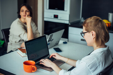 Blonde and brunette work on laptops, sitting at a table in the kitchen facing each other. Coworking in cozy home interior.