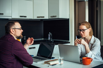 Home office during self-isolation. Husband and wife with glasses sitting at a table in the kitchen, remotely working together on laptops, discussing and smiling. Family freelance.