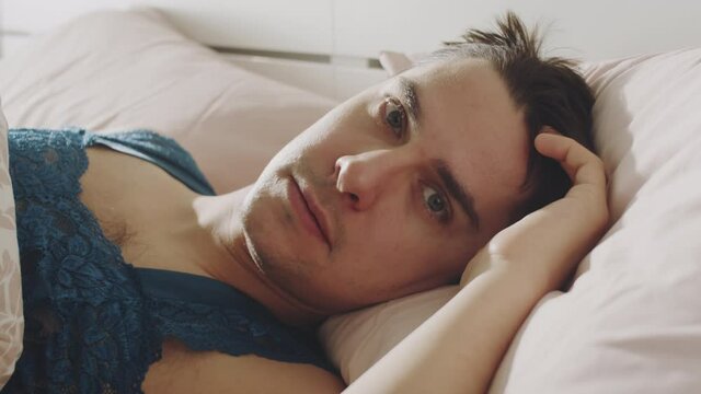 Portrait of young handsome transgender man in lace nightwear lying in bed and looking at camera after waking up in morning at home
