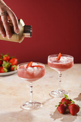 Female hand pouring cocktail our of shaker into a glass with red strawberry dessert with ice cubes and crashed ice and strawberry slices on burgundy background