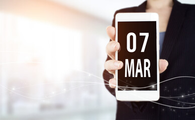 March 7th. Day 7 of month, Calendar date. White smartphone with Calendar date in businesswoman hand...