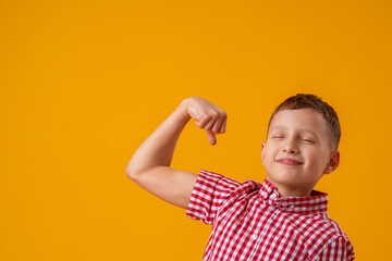 positive, confident 8-year-old boy in shirt smiles with his eyes closed and raises his hand into fist, flexing muscles in his arms, feeling strong, full of energy after healthy protein lunch, workout